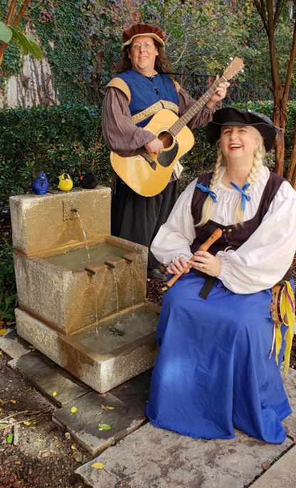 Fergus and Winifred posing by a small fountain. Winifred is sitting beside the fountain holding a recorder. Fergus is standing behind her with his guitar. They are both dressed in blue renaissance garb. Three plush birds are sitting on top of the fountain. 